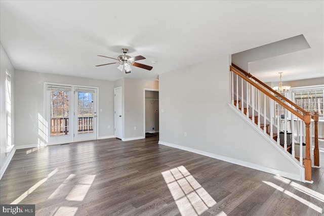 unfurnished living room featuring baseboards, dark wood-type flooring, stairs, and ceiling fan with notable chandelier