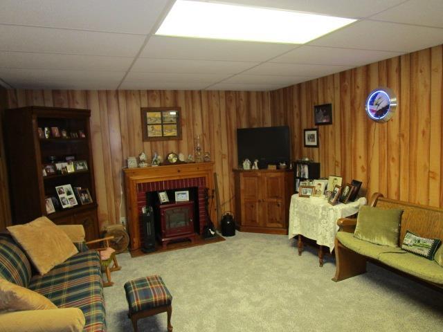 living area featuring a wood stove, wood walls, a drop ceiling, and carpet flooring