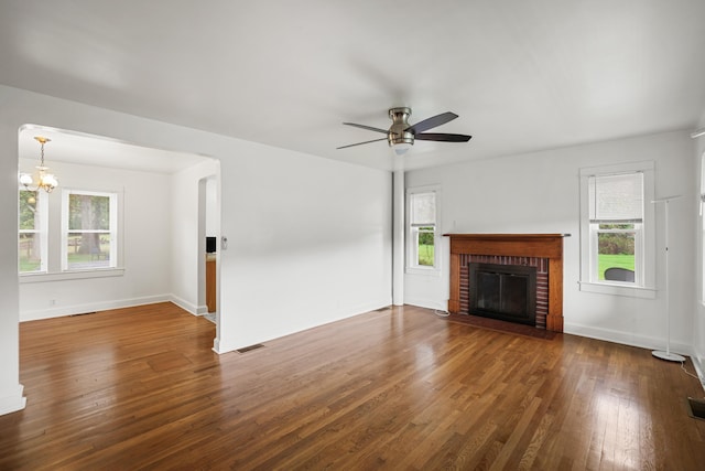 unfurnished living room featuring a healthy amount of sunlight, dark hardwood / wood-style flooring, ceiling fan with notable chandelier, and a brick fireplace