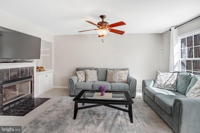 carpeted living room with baseboards, built in shelves, a ceiling fan, and a tiled fireplace