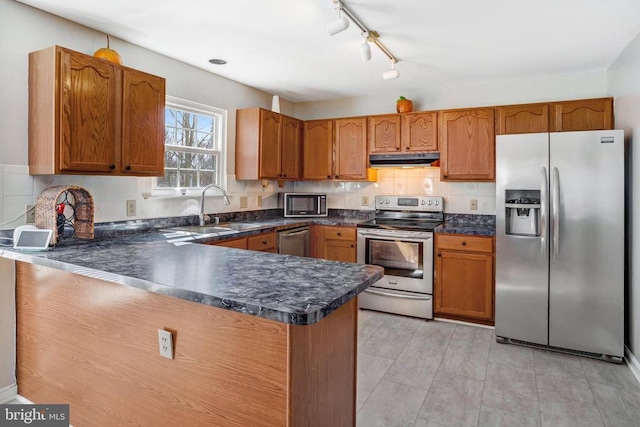 kitchen featuring a sink, under cabinet range hood, dark countertops, appliances with stainless steel finishes, and a peninsula