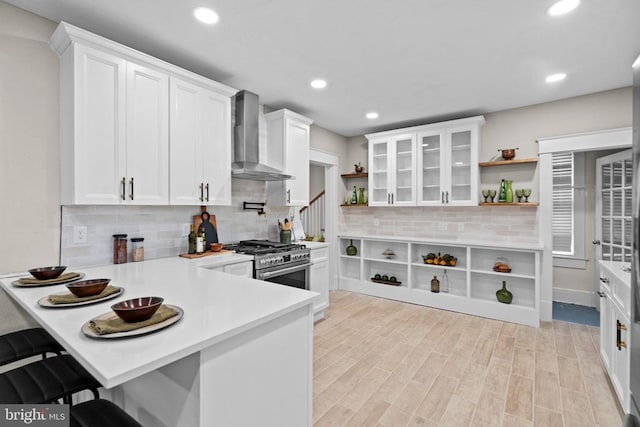 kitchen featuring wall chimney range hood, light hardwood / wood-style flooring, white cabinetry, a kitchen breakfast bar, and high end stove