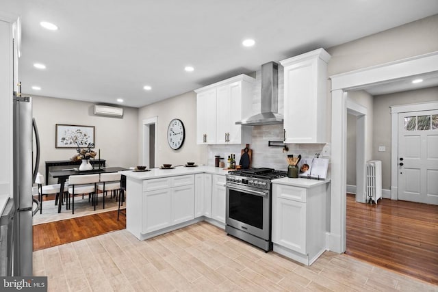 kitchen with a wall mounted AC, white cabinets, kitchen peninsula, stainless steel appliances, and wall chimney exhaust hood