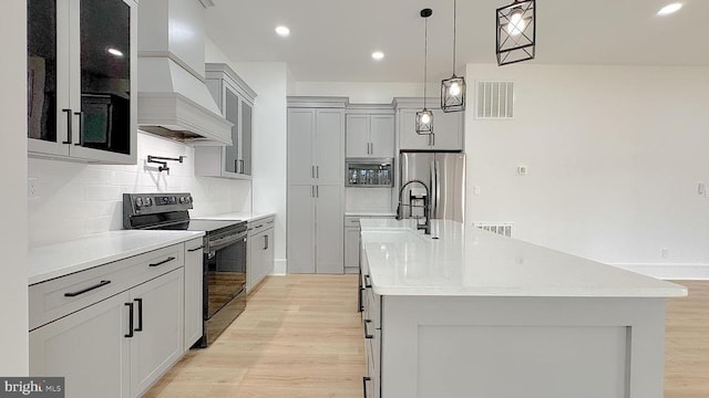 kitchen featuring appliances with stainless steel finishes, a kitchen island with sink, custom range hood, pendant lighting, and light hardwood / wood-style floors