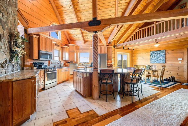 kitchen featuring appliances with stainless steel finishes, wood walls, beamed ceiling, a kitchen island with sink, and wood ceiling