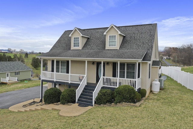 new england style home featuring covered porch, roof with shingles, fence, a front yard, and stairway