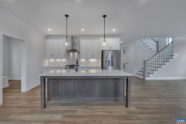 kitchen featuring baseboards, stainless steel fridge, wall chimney range hood, light countertops, and an island with sink