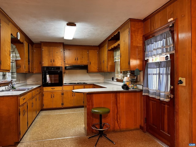 kitchen with a peninsula, brown cabinets, a sink, oven, and under cabinet range hood