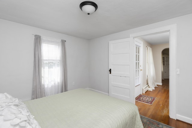 bedroom featuring arched walkways, wood finished floors, baseboards, and a baseboard heating unit