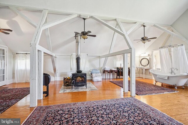 living room featuring a ceiling fan, a wood stove, high vaulted ceiling, wood finished floors, and beam ceiling