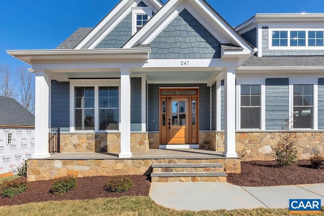 view of exterior entry featuring covered porch, roof with shingles, and stone siding