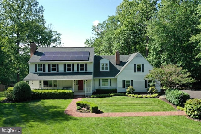 - view of front facade featuring a chimney, covered porch, solar panels, and a front yard