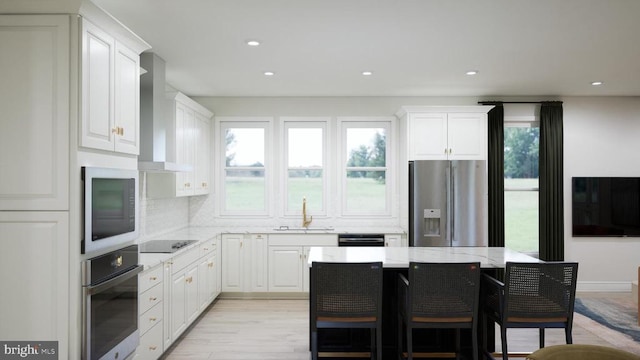  - kitchen featuring stainless steel appliances, wall chimney exhaust hood, white cabinets, light stone counters, and a kitchen bar