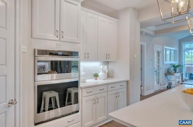 kitchen featuring an inviting chandelier, stainless steel double oven, white cabinets, light countertops, and decorative backsplash