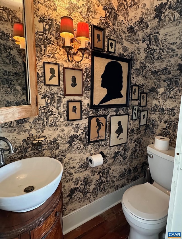 half bathroom with wood finished floors, wallpapered walls, toilet, vanity, and baseboards