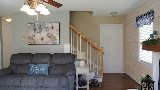 living room featuring dark wood-type flooring, ceiling fan, wooden walls, baseboards, and stairs