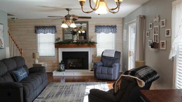 living room featuring a fireplace with raised hearth, wooden walls, ceiling fan with notable chandelier, wood finished floors, and stairs