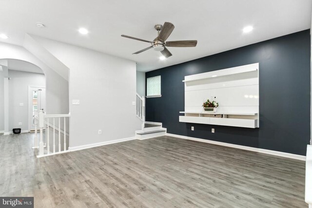 unfurnished living room with wood finished floors, arched walkways, a ceiling fan, recessed lighting, and baseboards