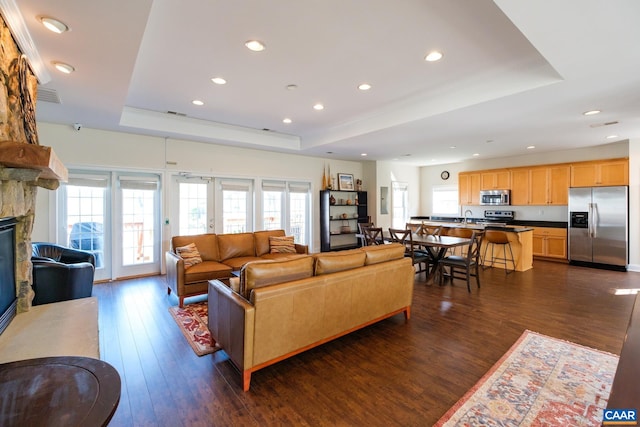 living room with a raised ceiling, a stone fireplace, recessed lighting, and dark wood-type flooring