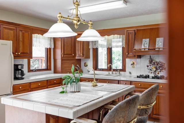 kitchen featuring freestanding refrigerator, brown cabinets, tile countertops, and a kitchen island with sink