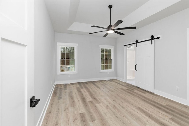 unfurnished bedroom featuring ceiling fan, a raised ceiling, a barn door, ensuite bath, and light hardwood / wood-style flooring