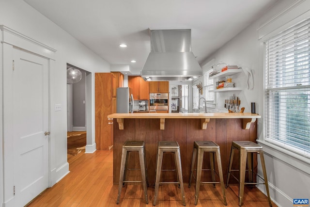 kitchen with brown cabinetry, a sink, island range hood, light wood-style floors, and stainless steel appliances