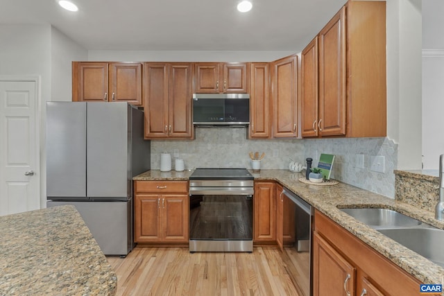 kitchen with a sink, stainless steel appliances, brown cabinets, light stone counters, and light wood-style flooring