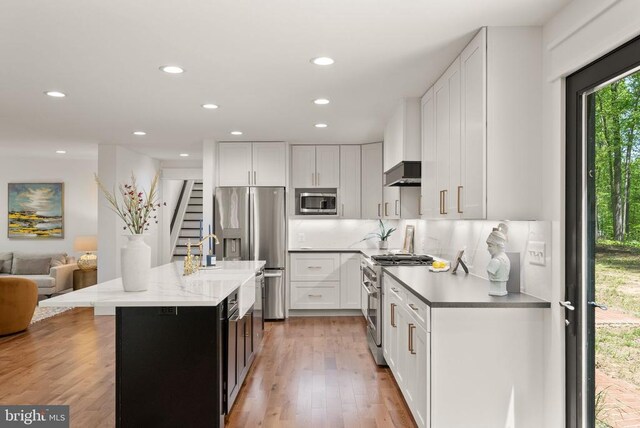  - kitchen featuring wall chimney exhaust hood, white cabinets, a kitchen island with sink, appliances with stainless steel finishes, and recessed lighting