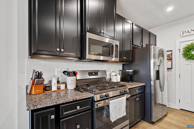 kitchen featuring stainless steel appliances, dark cabinetry, tasteful backsplash, light wood finished floors, and stone countertops