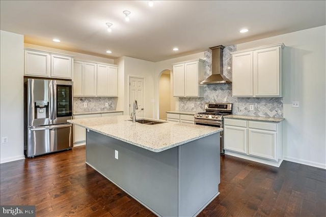 kitchen featuring a sink, a kitchen island with sink, wall chimney exhaust hood, arched walkways, and appliances with stainless steel finishes