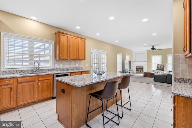 kitchen with a sink, stainless steel dishwasher, a glass covered fireplace, a ceiling fan, and brown cabinetry