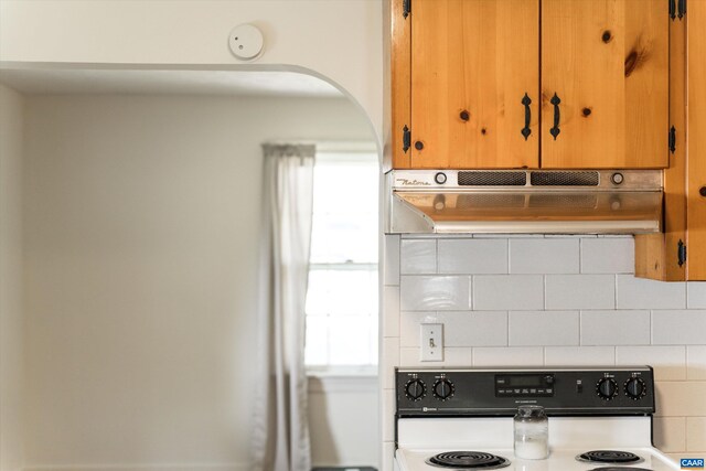 kitchen with brown cabinetry, tasteful backsplash, under cabinet range hood, arched walkways, and stove