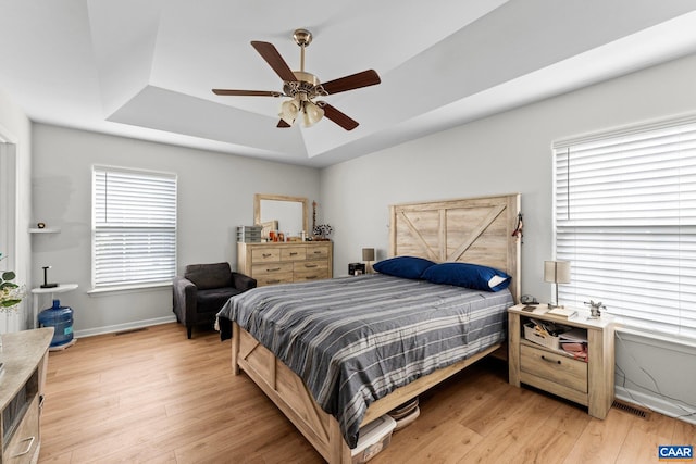 bedroom featuring visible vents, a ceiling fan, a tray ceiling, baseboards, and light wood-style floors