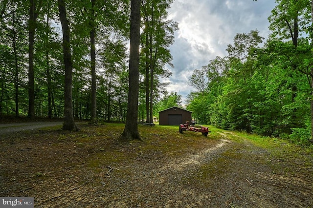 view of yard with a detached garage, dirt driveway, an outbuilding, and a view of trees