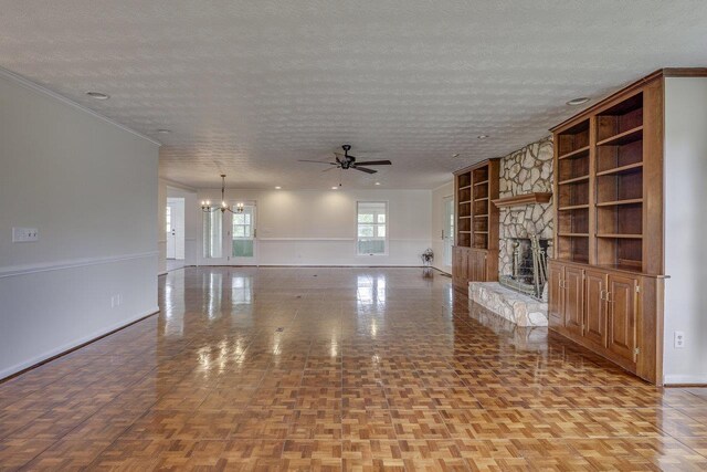 unfurnished living room featuring ceiling fan with notable chandelier, a textured ceiling, built in shelves, baseboards, and a stone fireplace
