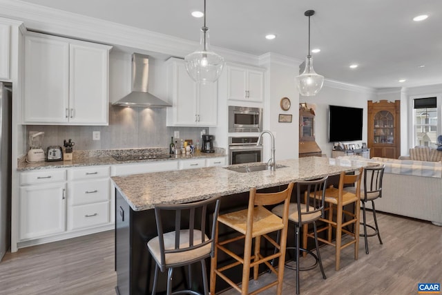 kitchen with wall chimney range hood, open floor plan, appliances with stainless steel finishes, a sink, and wood finished floors