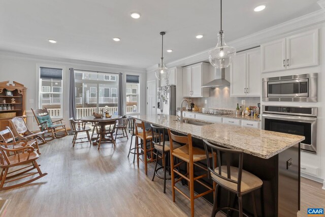 kitchen featuring stainless steel appliances, a sink, wall chimney range hood, crown molding, and white cabinetry
