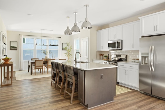 kitchen featuring wood finished floors, a center island with sink, a sink, appliances with stainless steel finishes, and backsplash