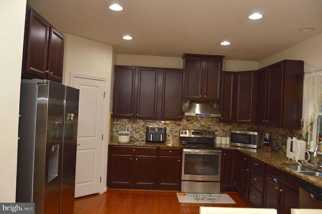 kitchen with appliances with stainless steel finishes, under cabinet range hood, a sink, backsplash, and dark stone countertops