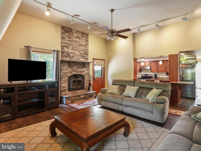  - living room featuring track lighting, a towering ceiling, dark wood-style floors, a ceiling fan, and a stone fireplace