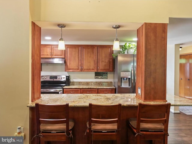  - kitchen with stainless steel appliances, under cabinet range hood, brown cabinetry, light stone counters, and hanging light fixtures