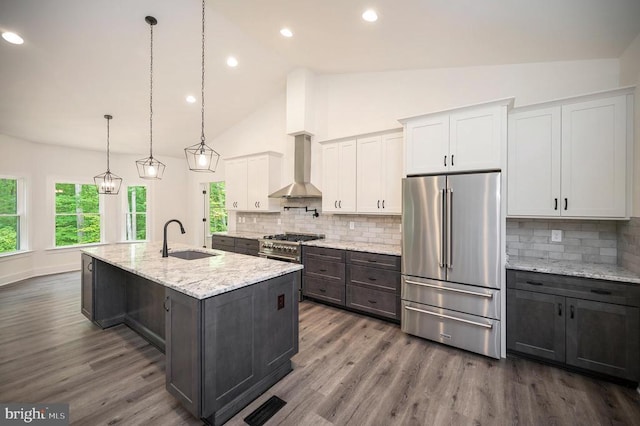  - kitchen with high end appliances, wall chimney exhaust hood, white cabinets, an island with sink, and light stone counters