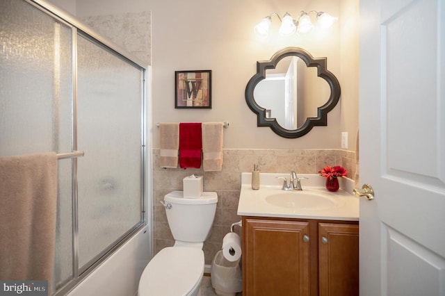 full bath featuring vanity, toilet, bath / shower combo with glass door, wainscoting, and tile walls
