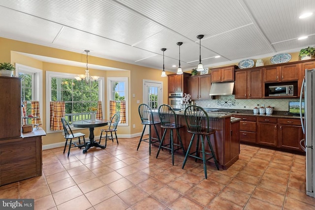  - kitchen featuring appliances with stainless steel finishes, under cabinet range hood, decorative backsplash, a kitchen breakfast bar, and light tile patterned floors
