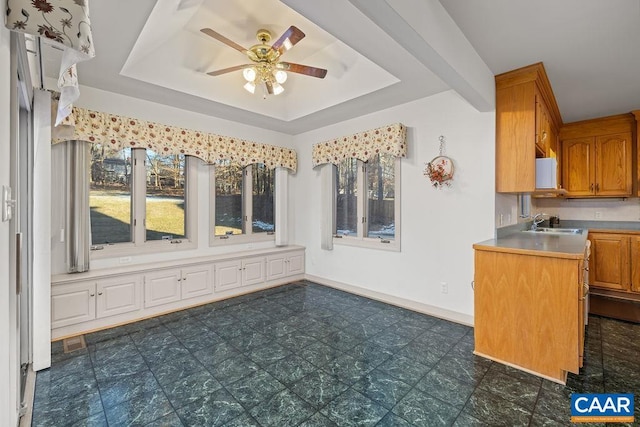 kitchen with a ceiling fan, a sink, a tray ceiling, brown cabinetry, and baseboards