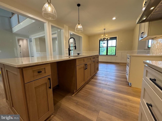  - kitchen with range hood, light wood-style floors, wainscoting, decorative light fixtures, and a center island with sink