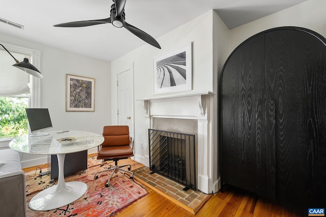 home office featuring a fireplace, a ceiling fan, and wood finished floors