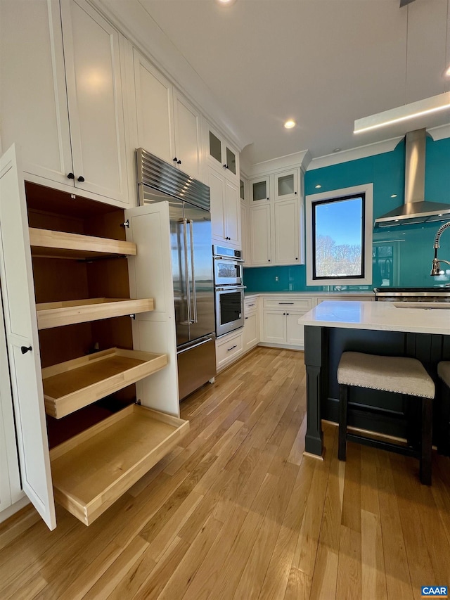 kitchen with light wood finished floors, wall chimney exhaust hood, stainless steel appliances, white cabinetry, and light countertops