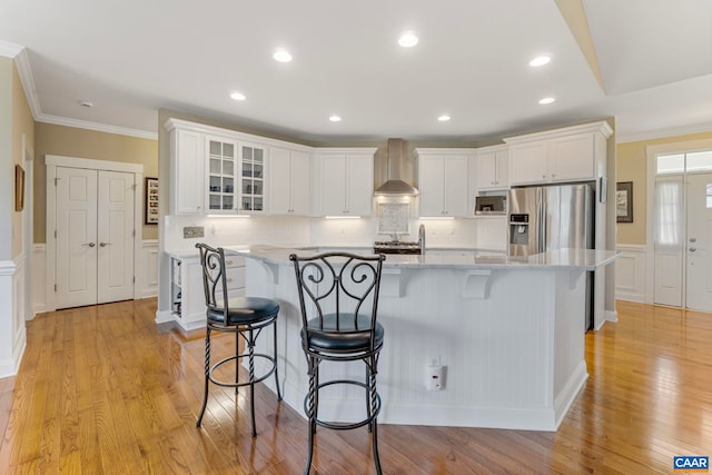 kitchen with stainless steel appliances, wall chimney range hood, ornamental molding, white cabinets, and a kitchen bar