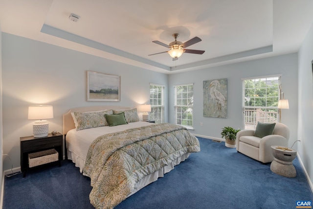 bedroom with ceiling fan, a raised ceiling, dark carpet, and multiple windows